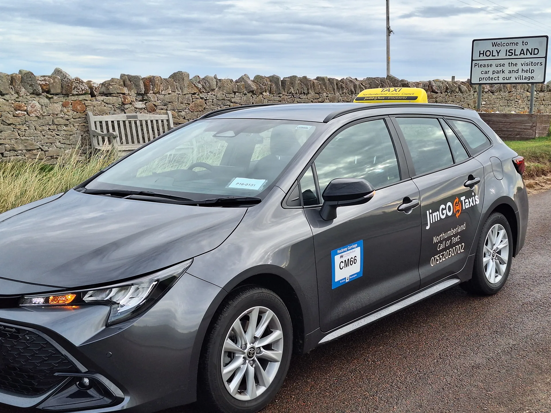 The JimGO Taxis Toyota Corolla Hybrid beside the Welcome to Holy Island sign