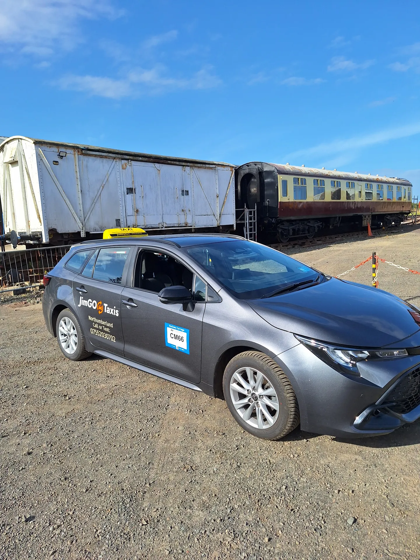 The JimGO Taxis Toyota Corolla Hybrid parked beside old railway carriages