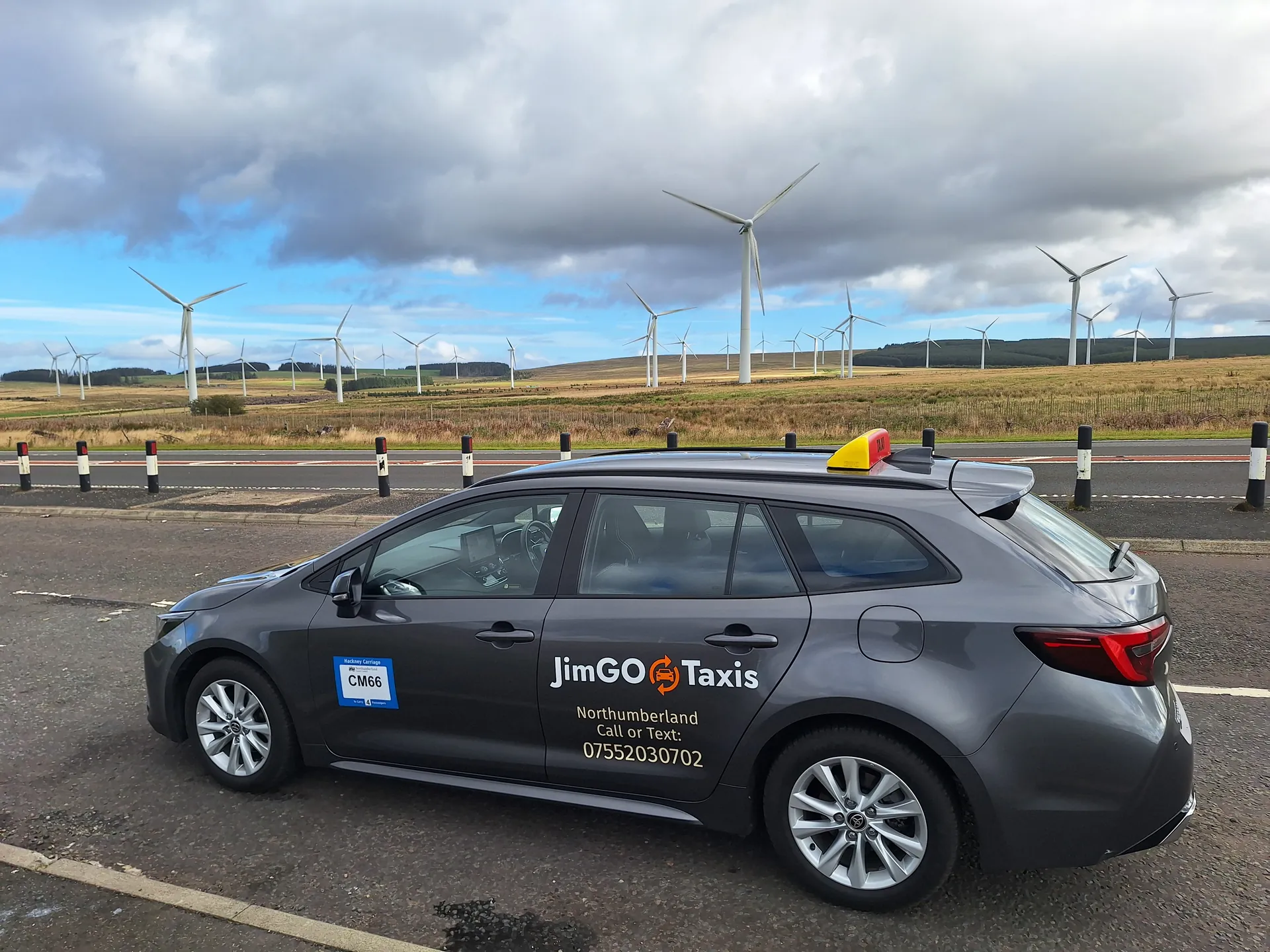 The JimGO Taxis Toyota Corolla Hybrid on a long road past a wind farm